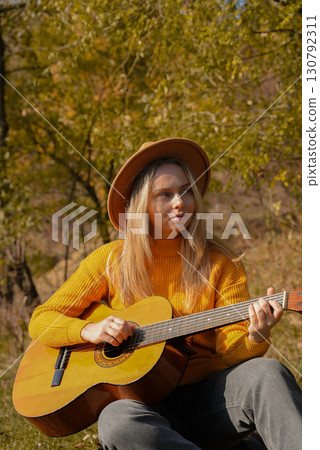 Blonde woman playing string guitar outdoors in autumn forest. Concept of sound therapy, mental health and wellness rituals. Calmness tranquility audio-sensory practices. Aura farming energy Blonde woman playing string guitar outdoors in autumn forest. Concept of sound therapy, mental health and wellness rituals. Calmness tranquility audio-sensory practices. Aura farming energy 130792311