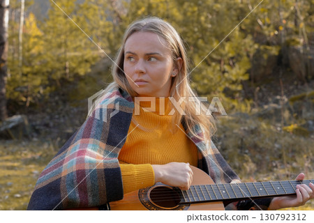 Portrait of smiling caucasian artist plays wooden acoustic guitar in autumnal park. Young songwriter plays string musical instrument hobby outside in nature fall time. Audio music healing Portrait of smiling caucasian artist plays wooden acoustic guitar in autumnal park. Young songwriter plays string musical instrument hobby outside in nature fall time. Audio music healing 130792312