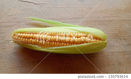 A single ear of ripe corn with its green husk partially peeled back, showcasing vibrant yellow kernels on a rustic wooden background. Captured in a top down view A single ear of ripe corn with its green husk partially peeled back, showcasing vibrant yellow kernels on a rustic wooden background. Captured in a top down view 130793614