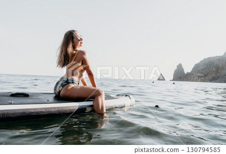 Woman Paddle Board Ocean - A woman in a bikini sits on a paddle board in the ocean, facing the water. Woman Paddle Board Ocean - A woman in a bikini sits on a paddle board in the ocean, facing the water. 130794585