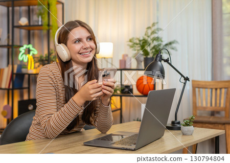Young woman in headphones at laptop holds up smartphone chats on social media messages happy smiling Young woman in headphones at laptop holds up smartphone chats on social media messages happy smiling 130794804