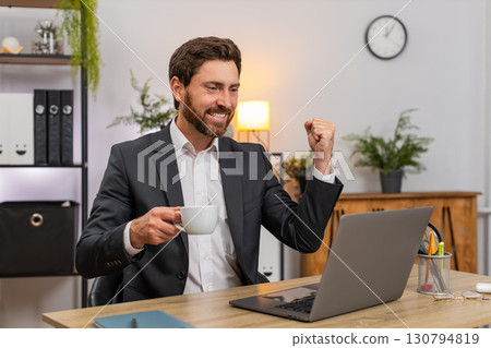Middle-aged businessman closes laptop raises espresso shot toasts success leans back chair relaxed Middle-aged businessman closes laptop raises espresso shot toasts success leans back chair relaxed 130794819