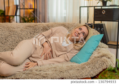 Adult woman holding stomach in pain on sofa at home, showing discomfort from food issues or cramps Adult woman holding stomach in pain on sofa at home, showing discomfort from food issues or cramps 130794933
