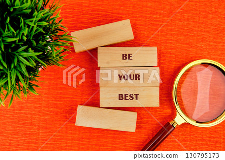 Copy space. Business concept. BE YOUR BEST assembled from wooden blocks in composition with magnifying glass and green plant on orange background, top view 130795173