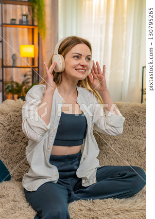 Adult woman dancing on home sofa with wireless headphones, enjoying favorite song energetic mood 130795265