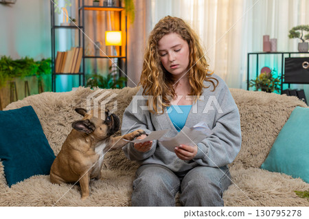 Caucasian woman looking worried at bills on home sofa while pug dog senses stress and stays close 130795278