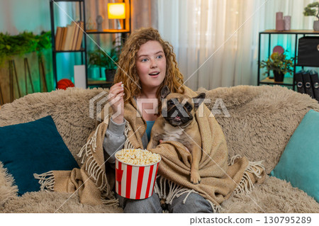Young woman watching TV on home sofa with pug dog under blanket, sharing popcorn and cozy evening Young woman watching TV on home sofa with pug dog under blanket, sharing popcorn and cozy evening 130795289