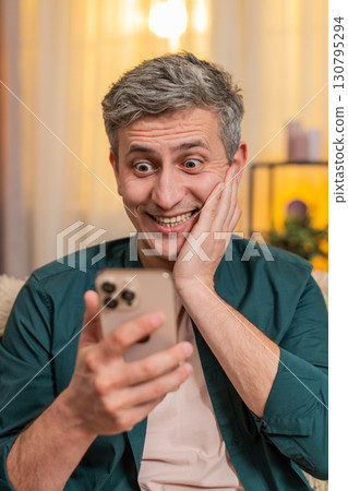 Young man reads smartphone message and reacts with open mouth and big eyes, showing surprise and joy 130795294