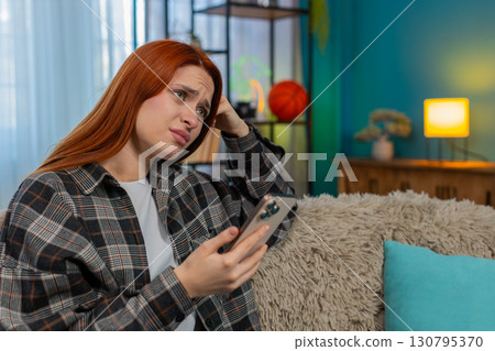 Woman sitting on sofa holding smartphone looking disappointed at screen after bad message or news Woman sitting on sofa holding smartphone looking disappointed at screen after bad message or news 130795370
