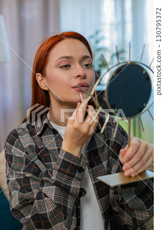 Young pretty woman applying makeup looking in mirror smiling while drawing lips with lipstick gently Young pretty woman applying makeup looking in mirror smiling while drawing lips with lipstick gently 130795372