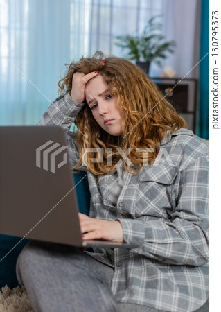 Young woman sits on sofa with laptop upset holding hand on forehead feeling failure and frustration Young woman sits on sofa with laptop upset holding hand on forehead feeling failure and frustration 130795373