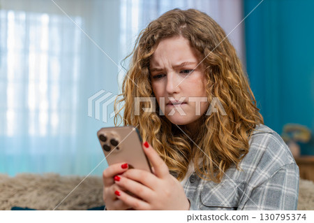 Young woman sits on sofa texting with anger waving hand annoyed by message using mobile phone chat 130795374