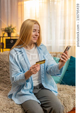 Young woman paying with credit card, smartphone transaction, relief, success at home couch, sunset 130795439