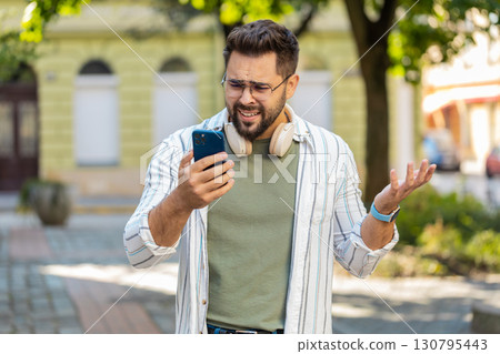 Upset young man using smartphone typing browsing, loses becoming shocked bad news on city street 130795443