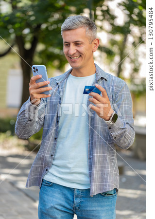 Happy mature man using credit bank card and smartphone purchases online shopping on city street 130795484