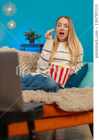 Caucasian woman enjoying movie on sofa eating popcorn laughing watching laptop indoors smiling happy Caucasian woman enjoying movie on sofa eating popcorn laughing watching laptop indoors smiling happy 130795513