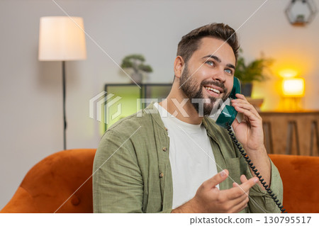 Smiling young man making wired telephone call conversation with friends sitting on sofa at home 130795517