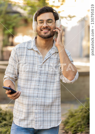 Happy Indian man in wireless headphones choosing, listening disco music on smartphone on street Happy Indian man in wireless headphones choosing, listening disco music on smartphone on street 130795819
