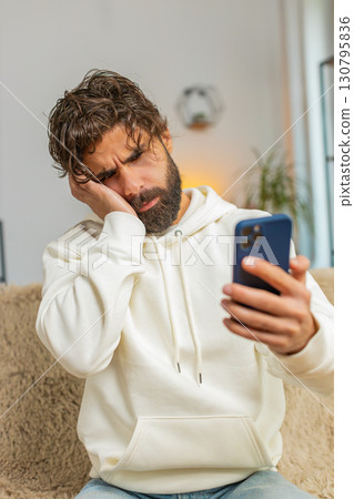 Upset displeased Indian man guy using smartphone reading bad news sitting on couch in living room Upset displeased Indian man guy using smartphone reading bad news sitting on couch in living room 130795836