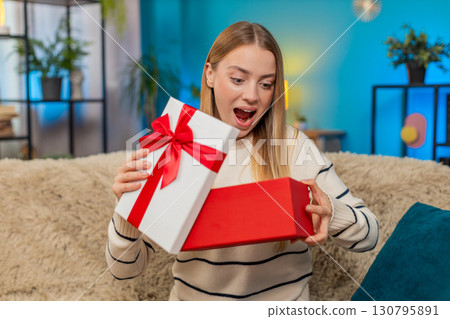 Caucasian woman opening birthday surprise gift on sofa smiling excited indoors happy joyful reaction Caucasian woman opening birthday surprise gift on sofa smiling excited indoors happy joyful reaction 130795891