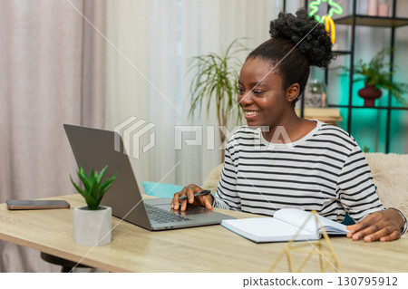 African American woman working focused on laptop sitting home on sofa writing notes in living room 130795912