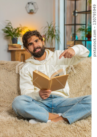 Indian man reading interesting book, turning pages smiling enjoying literature sitting on sofa 130795949
