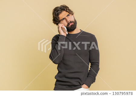 Disinterested young Indian man having annoyed boring talk on smartphone against beige background Disinterested young Indian man having annoyed boring talk on smartphone against beige background 130795978