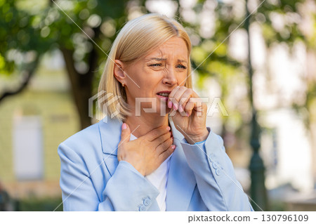 Sick mature businesswoman in formal suit suffering from sore throat while standing on city street 130796109