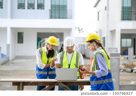 Group of contractors, engineers and formats in safety vests with helmets working with laptop, standing on under-construction building site. Home building project. Engineer foreman discusses with a 130796927
