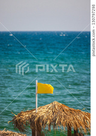 Yellow warning flag on a beach umbrella made of palm leaves, with turquoise sea waves in the background, symbolizing moderate swimming conditions. 130797298