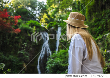 Young woman in a straw hat admiring a waterfall in a lush green garden during summer. 130797299
