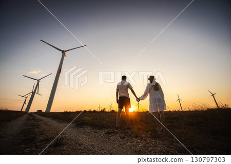 Couple holding hands walking near wind turbines at sunset, symbolizing love, harmony, and sustainable future. Couple holding hands walking near wind turbines at sunset, symbolizing love, harmony, and sustainable future. 130797303