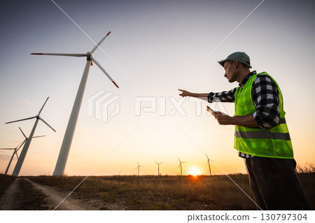 Engineer in safety vest pointing towards wind turbines at sunset, concept of renewable energy development and sustainable power control. 130797304