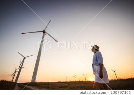 Middle-aged woman in hat walking near wind turbines at sunset, symbolizing eco-friendly lifestyle, renewable energy, and sustainable rural living. 130797305