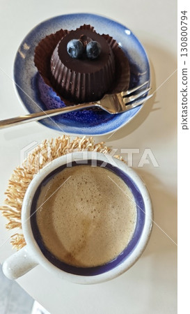 Cup of coffee with creamy foam and a small chocolate dessert topped with blueberries on a ceramic plate Cup of coffee with creamy foam and a small chocolate dessert topped with blueberries on a ceramic plate 130800794