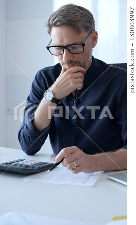 Businessman using a calculator and analyzing financial documents while preparing paperwork at an office desk. Audit and taxes, business concept 130803997