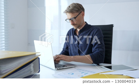Professional businessman wearing glasses, intensely working on laptop, typing amid modern office environment with tablet and documents surrounding workspace. Business people concept 130804019