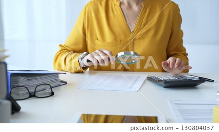 Female accountant wearing yellow shirt is examining a financial report with a magnifying glass while using a calculator and laptop in an office setting, close up. Audit and taxes in business 130804037