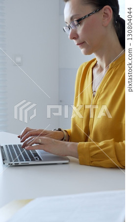 Businesswoman wearing yellow shirt sitting at white desk typing on laptop in bright modern office, working productively on computer. Business people concept Businesswoman wearing yellow shirt sitting at white desk typing on laptop in bright modern office, working productively on computer. Business people concept 130804044