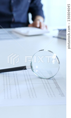 Magnifying glass highlighting a financial report with a businessman working on a laptop in the blurred background. Audit and taxes in business Magnifying glass highlighting a financial report with a businessman working on a laptop in the blurred background. Audit and taxes in business 130804045