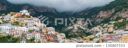 View of Positano's vibrant hillside buildings in white, yellow, orange, and red, surrounded by green vegetation, cliffs, and low hanging clouds. 130807333