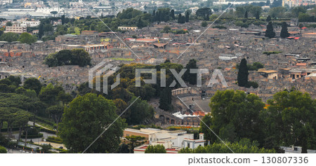 Aerial view of the ancient ruins of Pompeii in Italy, featuring the amphitheater, city layout, dense greenery, and modern urban areas in the distance. 130807336