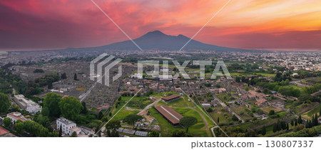 Aerial view of Pompeii's ruins with grid like streets, surrounded by greenery and modern buildings, with Mount Vesuvius under a fiery sunset sky. 130807337