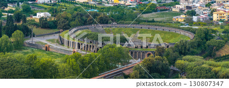 Aerial view of the oval shaped Roman amphitheater in Pompeii, Italy, surrounded by greenery, open spaces, and modern residential buildings. 130807347