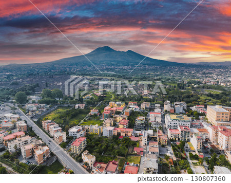 Aerial view of Pompei, Italy, featuring Mount Vesuvius under a colorful sunset. The scene includes ancient ruins, red tiled roofs, and green spaces. Aerial view of Pompei, Italy, featuring Mount Vesuvius under a colorful sunset. The scene includes ancient ruins, red tiled roofs, and green spaces. 130807360