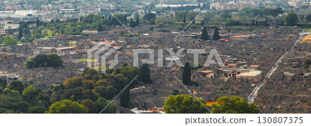 Aerial view of Pompeii's ancient ruins, featuring the amphitheater, stone streets, and buildings, with modern urban areas and greenery in the background. 130807375