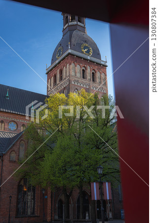 The Riga Cathedral stands with its Gothic architecture and clock tower, framed by a green tree, Latvian flags, a red element, and a bright blue sky. The Riga Cathedral stands with its Gothic architecture and clock tower, framed by a green tree, Latvian flags, a red element, and a bright blue sky. 130807384