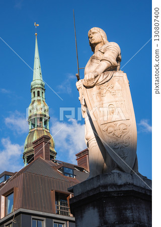 A statue of a historical figure with a sword and shield stands before the spire of St. Peter's Church in Riga, Latvia, under a clear blue sky. 130807400