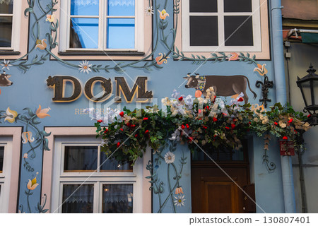 A soft blue building facade in Riga's old town features floral patterns, a whimsical cow illustration, gold DOM sign, and vibrant flower arrangements. A soft blue building facade in Riga's old town features floral patterns, a whimsical cow illustration, gold DOM sign, and vibrant flower arrangements. 130807401
