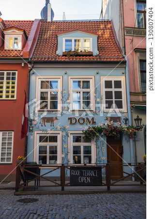 Light blue facade with floral and cat motifs, red tiled roof, cobblestone street, terrace sign, and Latvian flag in Riga's historic old town. 130807404